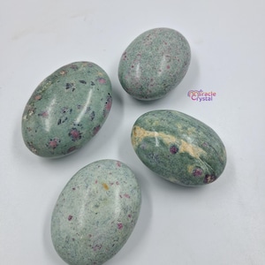 Może przedstawiać: Cztery owalne kamienie o nakrapianym wyglądzie. Kamienie są głównie w stonowanym zielonym kolorze, z małymi, nieregularnymi czerwonymi wtrąceniami. Kamienie są gładkie i wypolerowane i ułożone na białej powierzchni.