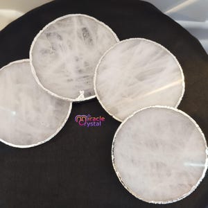 May include: Four circular, white crystal coasters with silver edges are shown on a black surface. The coasters have a natural, marbled appearance. The logo "Miracle Crystal" is visible.