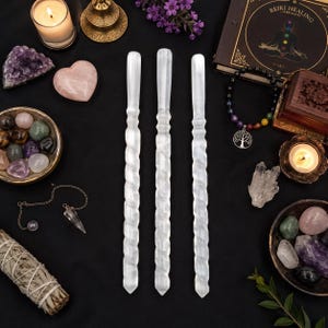 May include: Three white selenite wands with a spiral, twisted design arranged vertically in the centre. The wands are surrounded by various crystals, a chakra bracelet with a tree of life charm, a candle, a sage bundle, and a book on reiki healing.