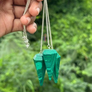 Może przedstawiać: Dwa zielone wahadełka z kryształu malachitu zawieszone na srebrnych łańcuszkach. Wahadełka mają fasetowany, spiczasty kształt. Łańcuszki trzymane są przez dłoń, na rozmytym zielonym tle.