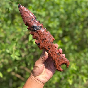 May include: A hand-carved, reddish-brown obsidian dagger with black inclusions. The dagger has a pointed tip and a jagged edge, with a handle featuring a curved design. The background is a blurred green foliage.
