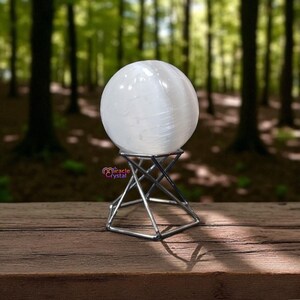 May include: A large, spherical, white selenite crystal ball sits on a silver metal stand. The stand has a geometric design. The background is a blurred view of a forest with tall trees. The crystal ball is the focal point.