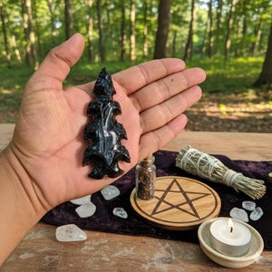 May include: A carved black obsidian arrowhead, held in a hand, with intricate details. The background includes a wooden table with a pentagram, a small candle, and a bundle of sage. Crystals and a small glass jar are also visible.