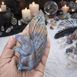 Puede incluir: Una figura de ardilla de piedra tallada en tonos azules, marrones y verdes. La ardilla está en posición sentada con la cola hacia arriba. La figura está detallada con pelo y rasgos tallados.
