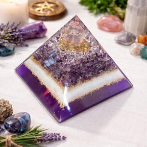 May include: A purple and clear resin pyramid with amethyst crystals, gold flakes, and a copper coil. The pyramid has a white band and is surrounded by other crystals and lavender sprigs.