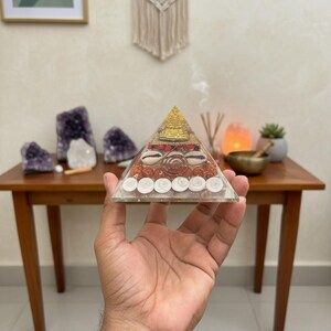 Puede incluir: Un dispositivo de orgón transparente en forma de pirámide con conchas, piedras y detalles dorados incrustados. La pirámide se sostiene en una mano, con una mesa de madera y objetos decorativos en el fondo, incluyendo cristales de amatista.