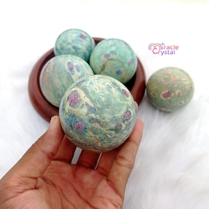 Puede incluir: Esferas de piedras preciosas en tonos turquesa, verde y rosa. Las esferas se muestran en un cuenco de madera y se sostienen en una mano. Las esferas tienen un aspecto veteado con inclusiones rosas y rojas. Diámetro aproximado de 5 cm.