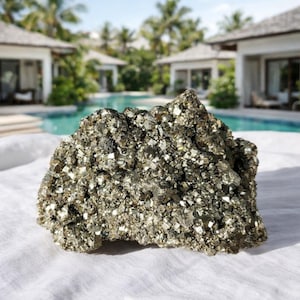 May include: A large, rough pyrite specimen with a metallic, gold-like luster. The natural stone is composed of many small, reflective crystals. The pyrite is displayed on a white surface, with a blurred background of a pool and tropical buildings.