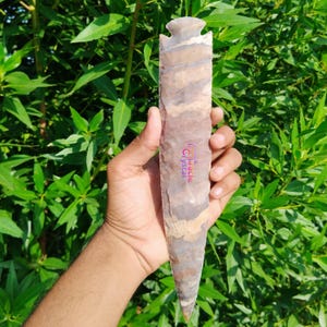 Puede incluir: Un objeto de piedra largo y tallado con un extremo puntiagudo y una parte superior con muescas, sostenido contra un fondo de follaje verde. La piedra presenta bandas de color marrón, gris y blanco. El texto "Miracle Crystal" es visible.