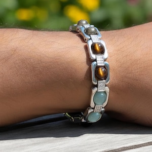 Puede incluir: Pulsera de eslabones plateados con ojo de tigre, piedras verde-azuladas y cristales transparentes. La pulsera se lleva en la muñeca, con un fondo verde borroso.