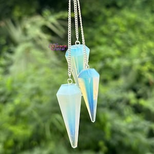 Puede incluir: Tres péndulos de cristal opalino colgando de cadenas plateadas. Cada péndulo tiene forma facetada de lágrima con tonos iridiscentes azules y amarillos. El fondo es verde borroso, lo que sugiere un entorno al aire libre. El logotipo de Miracle Crystal es visible.