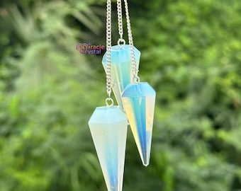 Op de afbeelding: Drie opalescente kristallen pendels hangen aan zilveren kettingen. Elke pendel heeft een facetvormige, druppelvormige vorm met iriserende blauwe en gele tinten. De achtergrond is een wazig groen, wat wijst op een buitenomgeving. Het Miracle Crystal-logo is zichtbaar.