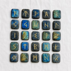 Può includere: Ventiquattro pietre runiche quadrate con simboli dorati. Le pietre sono un mix di blu, verde e nero con un aspetto maculato. Le rune sono disposte a griglia su una superficie bianca.