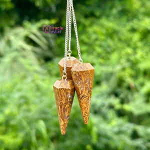 Puede incluir: Tres péndulos de piedras preciosas marrones y doradas con cadenas plateadas cuelgan sobre un fondo verde borroso. Los péndulos tienen forma de cono facetado. El texto "miracle crystal" es visible en la esquina superior izquierda.