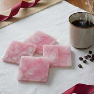 May include: Four square, pink coasters with a marbled appearance are arranged on a white cloth. A steaming mug of coffee and coffee beans are next to the coasters. A burgundy ribbon is draped across the scene.