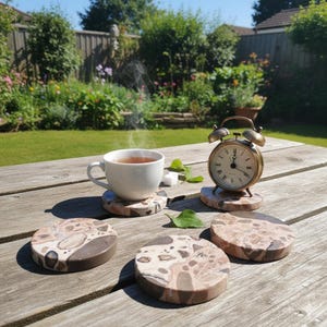 Pode incluir: Uma chávena branca com vapor, pousada sobre um descanso de copo estampado, ao lado de um despertador de latão vintage e vários descansos de copo de pedra. Os descansos de copo têm um padrão castanho e bege. A cena é ao ar livre, numa mesa de madeira.