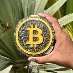 May include: A round, gold-colored Bitcoin symbol coaster. The Bitcoin symbol is in the center, surrounded by a gold border. The coaster is filled with small, dark gray stones. The background is a blurred view of green leaves.