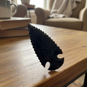 May include: A dark obsidian arrowhead with a notched design. The arrowhead is a deep black color and has a sharp, pointed tip. The arrowhead is resting on a wooden table.