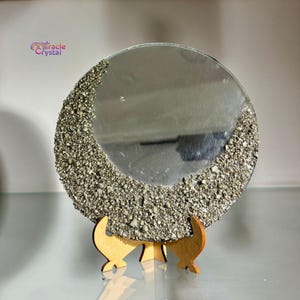 May include: A round mirror with a textured, gold-colored border, displayed on a wooden stand. The mirror's surface reflects the surrounding environment. The text "Miracle Crystal" is visible in the upper left corner.