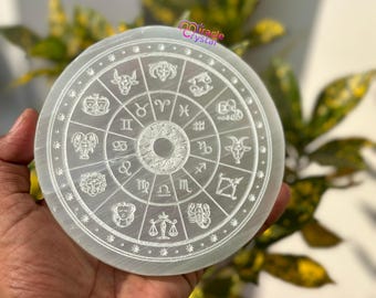 May include: A circular, white selenite crystal plate with engraved zodiac symbols. The plate has a central design and is encircled by the twelve astrological signs. The text "Miracle Crystal" is visible in the upper right corner.