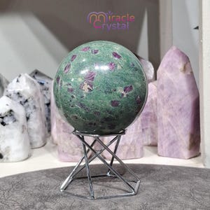 Könnte beinhalten: Eine grüne Kugel mit roten Einschlüssen, präsentiert auf einem silbernen Metallständer. Die Kugel befindet sich vor anderen Kristallen und dem Text "miracle crystal".