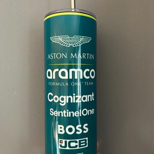 Puede incluir: Un vaso de color verde azulado del equipo Aston Martin Formula One con una pajita plateada. El vaso presenta el logotipo de Aston Martin y los logotipos de los patrocinadores, incluyendo Aramco, Cognizant, SentinelOne, BOSS, JCB y Valvoline.