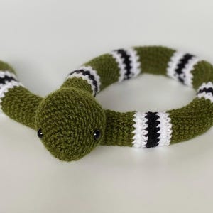 May include: A handmade, green crocheted snake toy with black and white striped details. The snake has a rounded head with small black eyes and is coiled. The toy is made of soft yarn, suitable for children.