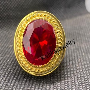 Könnte beinhalten: Ein goldfarbener Ring mit einem großen, oval geschliffenen, roten Edelstein. Der Ring hat einen strukturierten, seilartigen Rand um den Edelstein. Der Hintergrund ist eine strukturierte, graue Oberfläche.