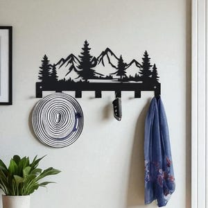 Op de afbeelding: Een zwarte metalen wandkapstok met een berg- en boom silhouet ontwerp. Een ingelijste print met het woord "WELCOME" hangt aan de muur. Een bank met een rieten mand en een potplant maken de entree decoratie compleet.