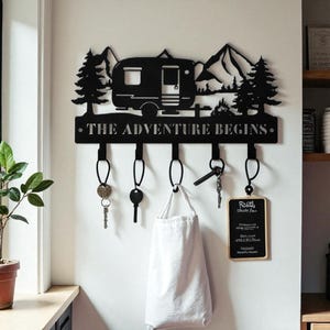 Op de afbeelding: Zwarte metalen sleutelhouder met een camperontwerp, bergen en bomen. De tekst "THE ADVENTURE BEGINS" is weergegeven. Inclusief vijf sleutelhaken en een klein schoolbord. Sleutels en een witte tas hangen eraan.