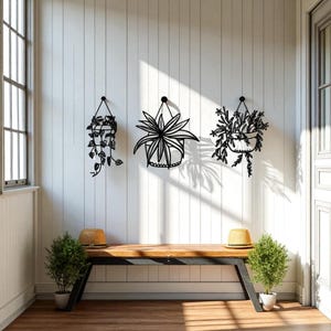 Peut inclure: Trois sculptures murales de plantes suspendues en métal noir sur un mur blanc. Les sculptures représentent divers motifs de plantes, notamment des vignes et des feuilles pointues. Un banc en bois et de petites plantes en pot sont en dessous.