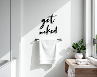 May include: A minimalist bathroom scene featuring a white towel hanging from a silver bar. Above the towel, the words "get naked" are displayed in black script. A small plant in a white pot sits on a wooden shelf.