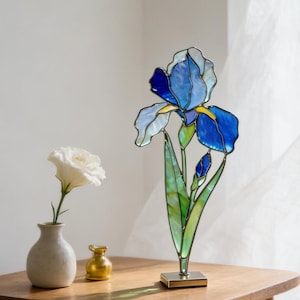 以下が含まれることがあります： 青と黄色の花びらと緑の葉を持つステンドグラスのアイリスの彫刻が、小さな木製のテーブルの上に立っています。小さな花瓶に入った白い花と金色の装飾品もテーブルの上にあります。銀色のフレームの時計が壁に掛かっています。