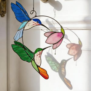 Puede incluir: Decoración de colibríes de vidriera con alas azules, verdes y naranjas, y una flor rosa. Los pájaros están suspendidos de un anillo de metal, colgado de un tirador de puerta. El diseño es colorido y detallado.