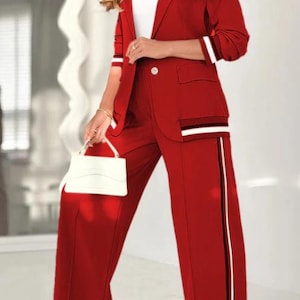 Puede incluir: Un traje pantal&oacute;n rojo con una blazer a juego. La blazer tiene detalles de rayas blancas y negras en las mangas y los bolsillos. El pantal&oacute;n tiene una raya vertical en el lateral. Se sostiene un bolso blanco en la mano.