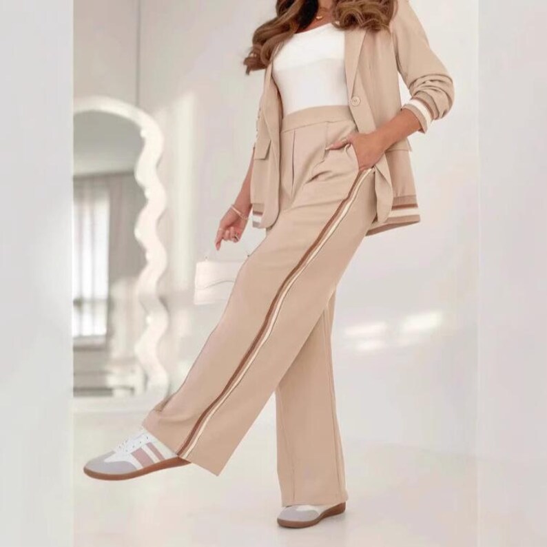 Puede incluir: Un traje pantal&oacute;n beige con una blazer a juego. La blazer tiene detalles a rayas blancas y marrones en las mangas. El pantal&oacute;n tiene una raya marr&oacute;n y blanca en el lateral. El conjunto se combina con zapatillas blancas y rosas.
