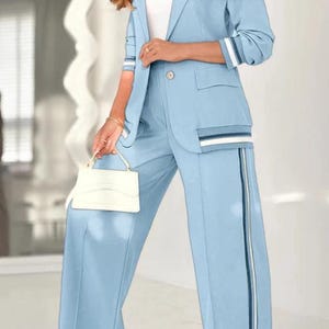 Puede incluir: Traje de pantal&oacute;n azul claro con una americana de un solo bot&oacute;n y pantalones de pernera ancha. La americana tiene detalles de rayas en las mangas y los bolsillos, y los pantalones tienen una raya vertical. Tambi&eacute;n se ve un peque&ntilde;o bolso de mano blanco.