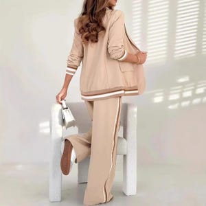 Puede incluir: Un traje de dos piezas beige con chaqueta y pantal&oacute;n de pernera ancha. La chaqueta tiene detalles de rayas blancas y marrones en las mangas y el dobladillo, con una raya a juego en el lateral del pantal&oacute;n. Se sostiene un peque&ntilde;o bolso blanco.