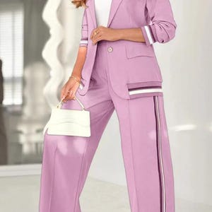 Puede incluir: Un traje pantal&oacute;n de color lila con una blazer con cierre de un solo bot&oacute;n, bolsillos y detalles a rayas en las mangas y los bolsillos. Los pantalones de pernera ancha tienen una raya vertical en el lateral. Un peque&ntilde;o bolso blanco completa el conjunto.