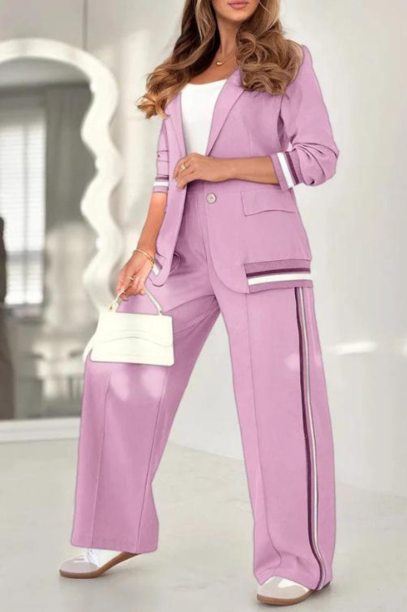 Puede incluir: Un traje pantal&oacute;n de color lila con una blazer con cierre de un solo bot&oacute;n, bolsillos y detalles a rayas en las mangas y los bolsillos. Los pantalones de pernera ancha tienen una raya vertical en el lateral. Un peque&ntilde;o bolso blanco completa el conjunto.