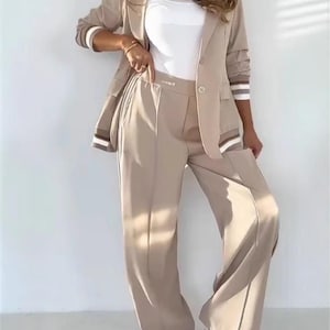 Puede incluir: Un traje pantal&oacute;n beige compuesto por una blazer y pantalones de pernera ancha. La blazer tiene un cierre de un solo bot&oacute;n y detalles a rayas en las mangas. Se lleva una camiseta blanca debajo. El conjunto se combina con zapatillas blancas y grises.