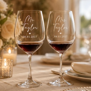 Puede incluir: Dos copas de vino de cristal transparente llenas de vino tinto. Cada copa presenta un texto grabado en blanco que dice Mr. Taylor o Mrs. Taylor, un pequeño icono de corazón y la fecha 06.01.2027. Las copas tienen tallos largos y finos.