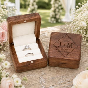 May include: A dark wood ring box with a white velvet interior holding two silver rings. The lid is engraved with a diamond-shaped border, leaf accents, and the text J + M, JESSICA, MICHAEL, and the date 07.18.2026 in a serif font.