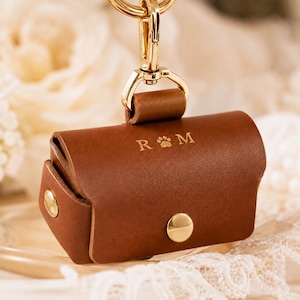 May include: A brown leather dog waste bag holder with gold-coloured clasp and hardware. The holder has a snap closure and features the initials "R & M" with a paw print symbol. Designed to attach to a leash or bag.