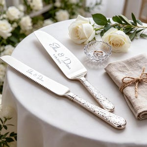 Puede incluir: Un juego de cuchillo y servidor de pastel plateado con nombres y fecha grabados. El juego presenta detalles florales ornamentados en los mangos. Dos anillos de boda están en un pequeño plato de vidrio, con rosas blancas y una servilleta de lino cerca.
