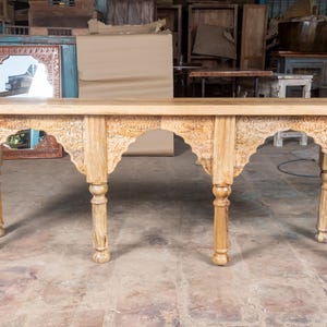 May include: A light-coloured wooden console table with intricate carved details. The table has a rectangular top and six arched legs with decorative carvings. The wood has a natural finish, showcasing the grain. The table is a piece of furniture.