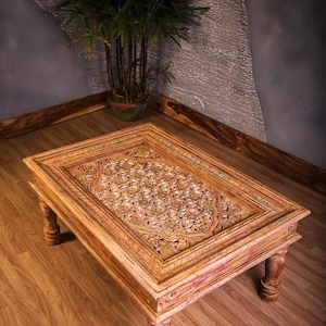 May include: Wooden coffee table with intricate floral carvings on the tabletop. The table has a rectangular shape with four sturdy legs. The wood has a warm, natural tone with hints of red. A potted plant is in the background.