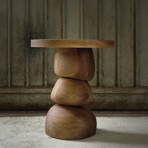 Puede incluir: Una mesa auxiliar de madera hecha a mano con un diseño apilado único. La mesa presenta una tapa redonda sostenida por tres segmentos de madera redondeados y de forma orgánica. La veta natural de la madera es visible, mostrando los cálidos tonos marrones. La mesa mide unos 51 cm de altura.