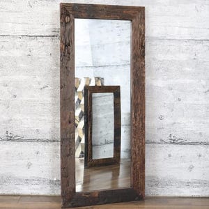 Pode incluir: Um espelho retangular grande com uma moldura de madeira castanha escura e rústica. A moldura tem uma aparência texturizada e desgastada. O espelho reflete um espelho menor e um objeto decorativo, contra uma parede cinza claro e branca.