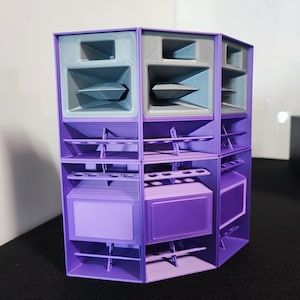 Puede incluir: Un sistema de altavoces modular en morado y gris, con múltiples compartimentos. El diseño es geométrico, con superficies y aberturas angulares. El sistema está diseñado para apilarse y organizarse en varias configuraciones.